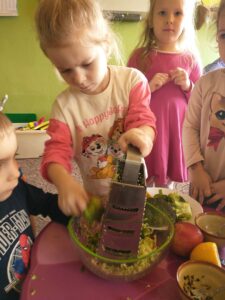 dziewczynka trze brokuła na tarce do warzyw, obok stoją inne dzieci, na stole leżą jabłko, cytryna, miseczki