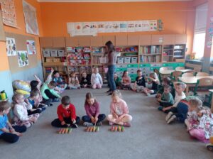 grupa dzieci siedzi w półkolu na dywanie, troje dzieci gra na instrumentach
