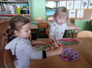 dwie dziewczynki siedzą prz stoliku, w dłoni trzymają pędzelek do malowania.