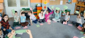 Dzieci siedzą na dywanie i podnoszą różową lub zieloną kartkę.