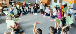 Dzieci siedzą na dywanie i podnoszą różową lub zieloną kartkę.