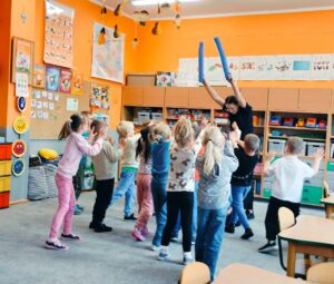 Dzieci wykonują gest oznaczający stop, trenerka trzyma nad nimi gąbkowe makarony