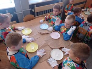 grupa dzieci siedzi przy stolikach, są ubrani w rękawiczki