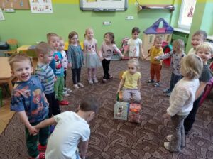 dzieci stoją w kole na dywanie, w środku siedzi dziewczynka, przed nią dwie torebki prezentowe