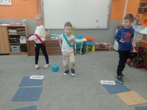 troje dzieci stoi na dywanie, jedno rzuca kostką