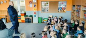 Dzieci siedzą na dywanie. Przed dziećmi stoi policjant