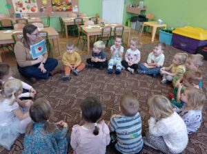 grupa dzieci siedzi z prowadzącą na dywanie i oglądają obrazki