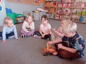 kilkoro dzieci siedzi na dywanie, chłopiec karmi misia