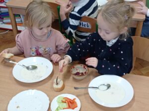 dwie dziewczynki siedzą przy stoliku, jedna smaruje kanapkę