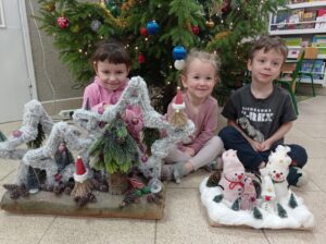 troje dzieci siedzi przed choinką, trzyma w ręku prace wykonane na konkurs