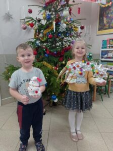 dwoje dzieci siedzi przed choinką, trzyma w ręku prace wykonane na konkurs