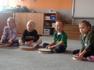 grupa dzieci siedzi na dywanie, przed nimi bębenki