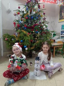 dwie dziewczynki siedzą przed choinką, trzyma w ręku prace wykonane na konkurs