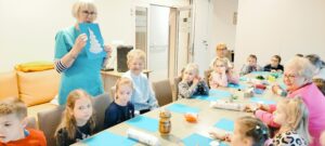 Dzieci i podopieczni siedzą przy stolikach. Pani tłumaczy na czym polega zadanie