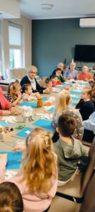Dzieci i podopieczni siedzą przy stolikach. Pani tłumaczy na czym polega zadanie