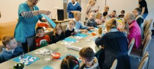 Dzieci i podopieczni siedzą przy stolikach. Pani tłumaczy na czym polega zadanie