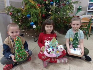 troje dzieci siedzi przed choinką, trzyma w ręku prace wykonane na konkurs