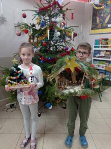 dwoje dzieci stoi przed choinką, trzyma w ręku prace wykonane na konkurs