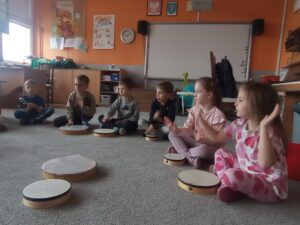 grupa dzieci siedzi na dywanie, przed nimi bębenki